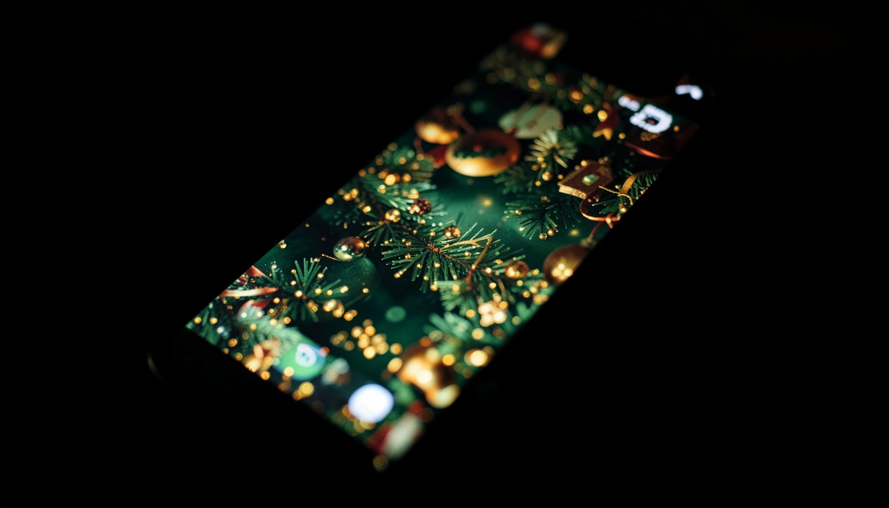 Dark, moody close-up photograph of a smartphone screen glowing with a festive Christmas wallpaper design, deep greens and warm reds against deep shadow